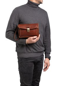 Borsa a tracolla in pelle marrone con pattina, chiusura in oro e tracolla removibile. Presenta bordi cuciti e forma rettangolare, tenuta da una persona.
