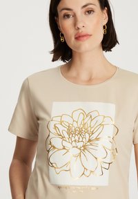 Beżowa koszulka z krótkim rękawem, ozdobiona złotym foliowym wzorem kwiatowym oraz tekstem pośrodku. Okrągły dekolt i gładka faktura materiału.