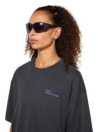 Kobieta z kręconymi włosami nosząca czarne okulary przeciwsłoneczne typu wraparound oraz ciemną, oversize'ową koszulkę z małym fioletowym napisem na piersi.