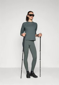 Athletisches Outfit mit einem gerippten, kurzen dunkelgrünen Shirt und passenden engen Leggings. Zubehör beinhaltet schwarze Wanderstöcke und Schuhe.