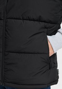 Schwarze Steppweste mit gestepptem Design, durchgehendem Reißverschluss und seitlicher Tasche. Der Stoff wirkt glatt und leicht. Etikett am Saum sichtbar.