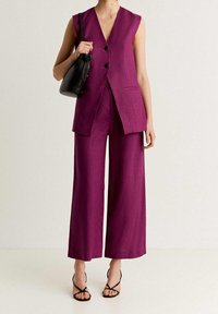 Fuchsia linnen gilet zonder mouwen en met twee knopen, gecombineerd met wijde broek in dezelfde kleur. Zwarte emmerzak en sandalen met banden.