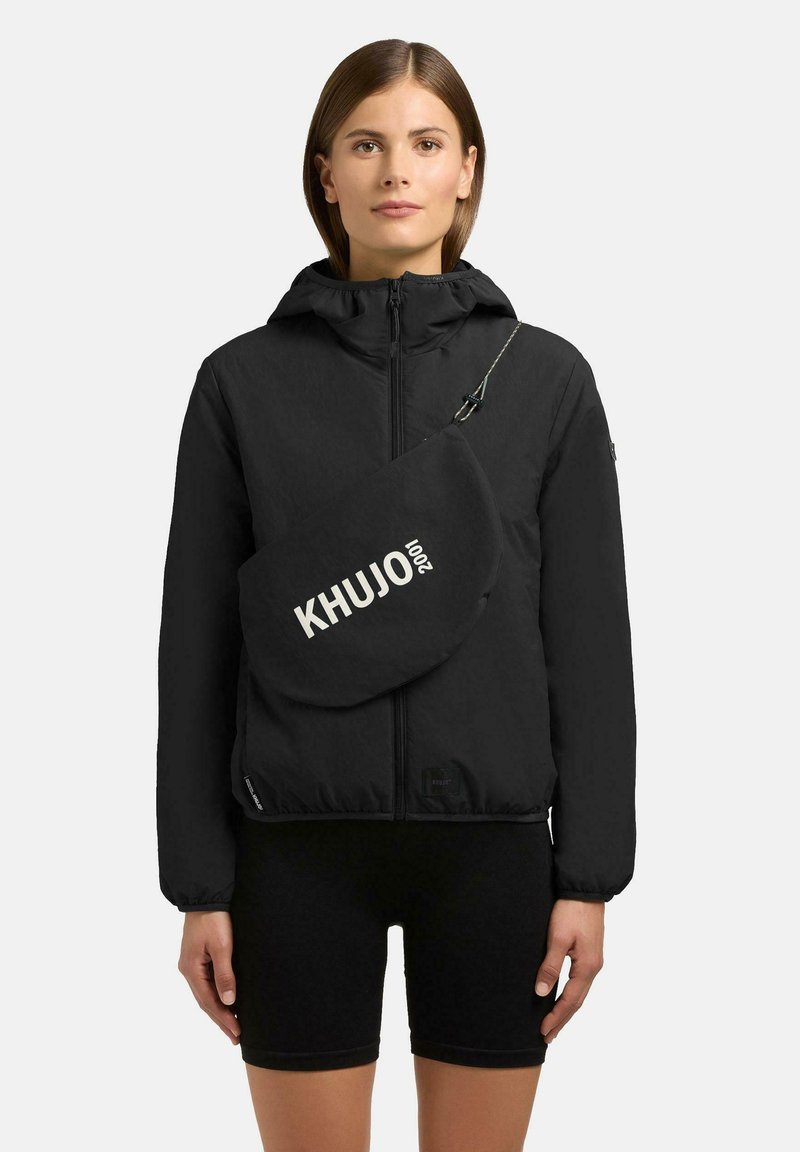 Kvinde iført en sort lynlåstrøje med hætte og en skuldertaske mærket "KHUJO 2001," kombineret med sorte cykelshorts.