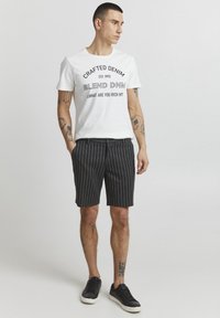 Maglietta bianca con testo grafico e logo neri, abbinata a pantaloni corti a righe scure. Sneakers nere completano l'outfit.