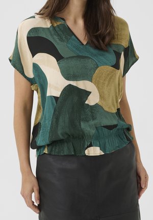 Frau trägt ein kurzärmeliges Oberteil mit abstraktem Muster in Grün-, Schwarz-, Beige- und Gelbtönen, kombiniert mit einem schwarzen Lederrock.