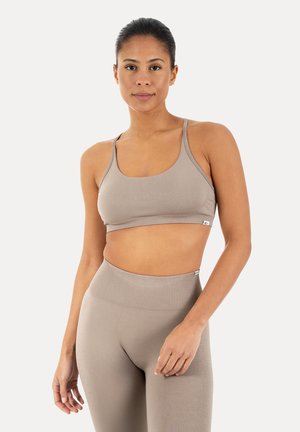 Kvinde stående iført beige sports-bh og højtaljede leggings, ser frem med neutralt udtryk, ensfarvet lys baggrund.