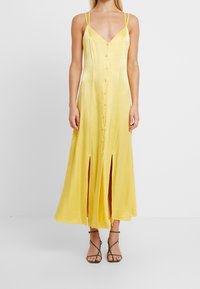 Robe maxi sans manches jaune avec un décolleté en V, des boutons à l'avant et des fentes hautes. Fabriquée en tissu doux avec un léger éclat.