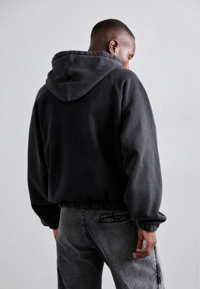 Gepachtete schwarze Kapuzenjacke mit einer Kordelzugkapuze und gerippten Bündchen. Getragen mit ausgewaschenen grauen Jeans, die ein distressed Muster zeigen.