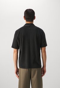 Lyhythihainen musta polo-paita rentoa leikkausta, jossa on litteä kaulus ja pehmeä materiaali; yhdistettynä väljiin vaaleanbeigeihin housuihin.