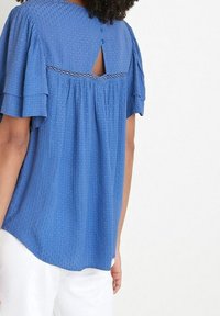 Modrá halenka s krátkým rukávem, texturovanými tečkami, detaily ve tvaru klíčové dírky na zádech a jemným lemem na švu. Měkká tkanina s volným střihem.
