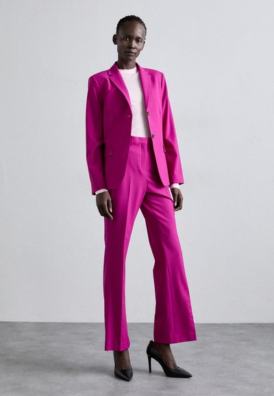 Kirkas vaaleanpunainen räätälöity puku, jossa on istuva bleiseri ja levenevät housut, yhdistettynä vaaleanpunaiseen puseroon ja mustiin korkokenkiin.