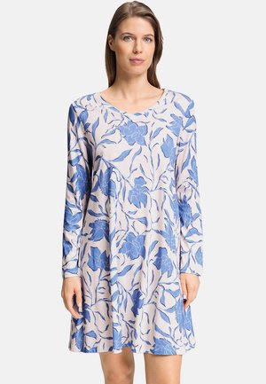 Frau trägt ein knielanges Kleid mit langen Ärmeln und blauem Blumenmuster auf weißem Hintergrund und steht vor einem schlichten Hintergrund.