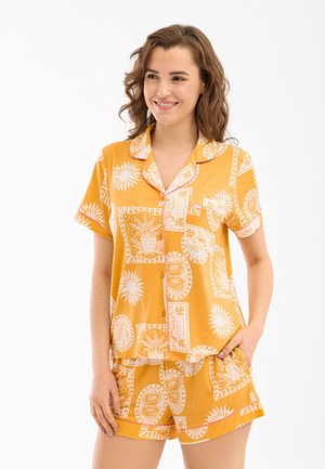 Nuori nainen hymyilee, hänellä on oranssi lyhythihainen nappipaita ja siihen sopivat shortsit, joissa on valkoisia aurinko- ja hedelmäkuvioita.