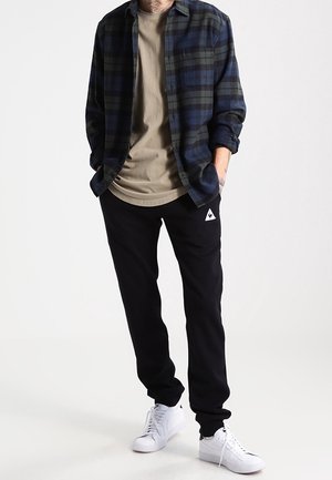 Rutet flannel skjorte i mørkeblå og grøn, beige t-shirt, sorte sweatpants med logo og hvide sneakers. Afslappet, lagdelt outfit.