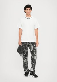 Camiseta blanca de manga corta con texto en el cuello negro, combinada con pantalones negros que presentan un estampado gráfico detallado y una gorra negra a juego.