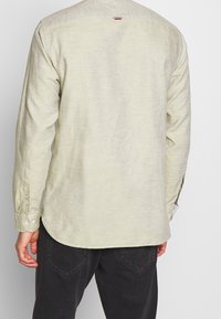 Muž v dlouhém světle béžovém tričku s dlouhým rukávem a tmavých kalhotách, zobrazený ze zadu na jednobarevném šedém pozadí.