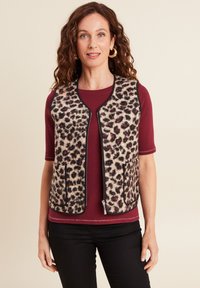 Beige luipaardprint vest met zwarte details, voorzien van twee voorzakken en een ritsluiting. Draagt over een mahoniekleurige top, tegen een neutrale achtergrond.