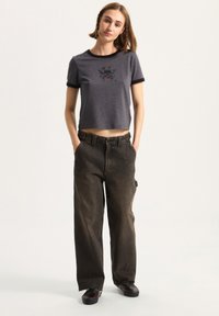 Top corto gris con ribete negro y un diseño gráfico, combinado con unos vaqueros oscuros de corte holgado. La modelo está de pie con las manos en los bolsillos, usando zapatos oscuros.