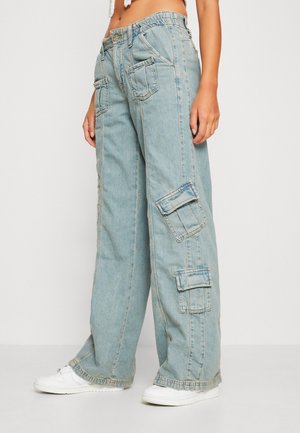 Lyseblå løsthængende denim cargobukser med flere klaplommer, båret med hvide sneakers, vist fra taljen og ned på en stående person.