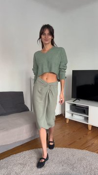 Hellgrüner, gekürzter Pullover, hochtaillierter, geknoteter, hellgrüner Rock und schwarze Mary Jane-Schuhe. Weiche Texturen und lässige Passform.