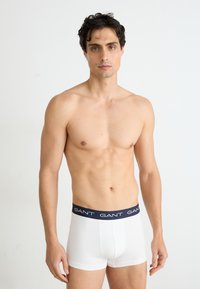 Weiße Boxershorts mit einem marineblauen Bund, auf dem der Markenname "GANT" in weißer Schrift steht. Glattes Material mit einer taillierten Passform.