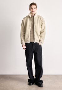 Bež flis jakna s visokim ovratnikom, patentnim zatvaračem sprijeda i rebrastim manžetama, uparen s trapericama tamne boje širokog kroja i crnim cipelama.