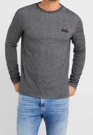 Tmavě šedé tričko s dlouhým rukávem, kulatým výstřihem, jemným logem značky na hrudi a strukturovanou látkou. Doplňuje jej světlé modré džíny.