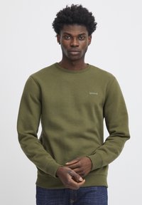 Jonge man met kort krullend haar, een olijfgroen sweatshirt met het "blend" logo aan, staand met zijn handen voor zich gekruist op een plain witte achtergrond.