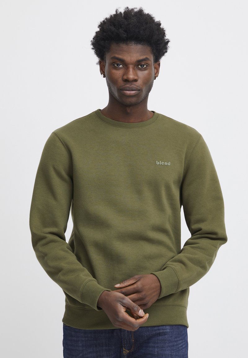 Joven con cabello rizado corto vistiendo una sudadera verde oliva con el logo "blend", de pie con las manos entrelazadas al frente sobre un fondo blanco liso.