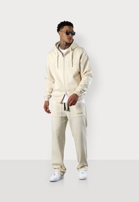 Hoodie zippé beige avec logo sur la poitrine, associé à un pantalon de survêtement assorti. Tissu lisse, coupe décontractée et baskets blanches complètent le look.