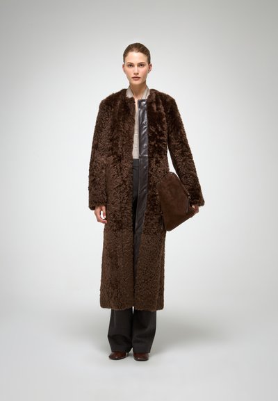 Bruin textuur bonten jas met een lange silhouette, voorzien van een glad leren voorkant en bijpassende bruine clutch. Model draagt grijze broek.