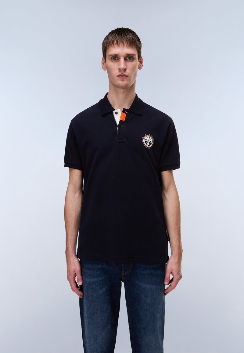 Μπλε μπλούζα πόλο με μίνιμαλ γιακά σε κρεμ χρώμα που δημιουργεί αντίθεση, πορτοκαλί λεπτομέρεια και κεντημένο λογότυπο στο στήθος. Συνδυάζεται με μπλε τζιν παντελόνι.