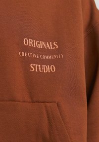 Bruine sweatshirt met de opdruk "ORIGINALS" en "STUDIO" in contrasterende tekst. Inclusief een voork pocket en zachte, gestructureerde stof.