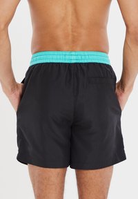 Zwarte zwemshorts met een turquoise elastische tailleband, uitgerust met zijzakken en een detail van een achterzak. Glad materiaal met een lichtgewicht ontwerp.