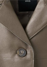 Gros plan d'une veste en cuir taupe avec un large col à revers et un seul grand bouton foncé, montrant la texture et les coutures détaillées.