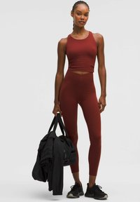 Vörösesbarna sportos crop top és hozzá illő, magas derekú legginsz. A modell egy fekete táskát tart, fekete sportcipőt visel. Sima textúra.