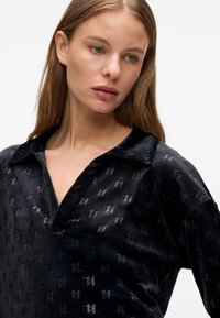 Camicia nera in velluto con colletto, motivi logo discreti e vestibilità morbida con texture liscia.