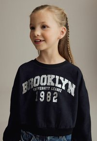 Sweatshirt court noir avec "BROOKLYN UNIVERSITY LEAGUE 1982" en lettres blanches texturées. Col et poignets côtelés.