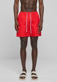 Pantaloni scurți de înot roșii, cu un brâu elastic și șnur alb, textură netedă și buzunare laterale. Purtați cu sandale de culoare deschisă.