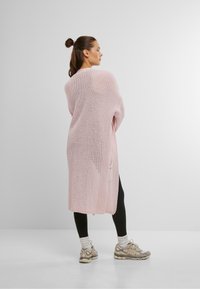 Lichtroze gebreide cardigan met een open ontwerp, lange mouwen en zijzakken, gedragen over zwarte leggings en lichtgekleurde sneakers.