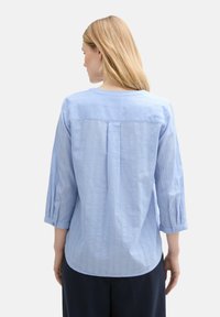 Light blue, textured shirt with three-quarter sleeves, a rounded neckline, and a back pleat detail. Fabric appears soft and breathable.