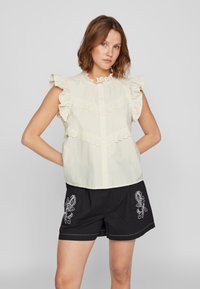 Kremfarget, kortermet bluse med volanger, blonder og knapper. Kombinert med svarte shorts med hvitt broderi.