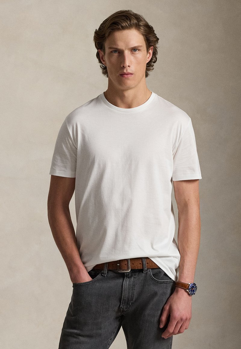 Νέος άνδρας με καστανά κατσαρά μαλ λιά, που φοράει λευκό κοντομάνικο μπλουζάκι, σκουρόχρωμο τζιν, καφέ ζώνη και ρολόι με μπλε καντράν και καφέ λουράκι, όρθιος.