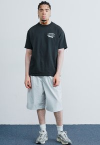 T-shirt en coton noir avec le logo « OBEY » et un short gris clair à mi-genoux. Le modèle porte des baskets grises avec des accents à motifs.