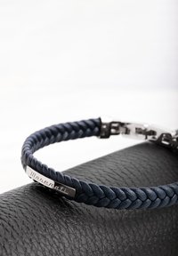 Mjärteblå läderarmband med flätad design, silverfäste och en metalldekorplatta präglad med en logotyp. Texturerad svart yta nedanför.