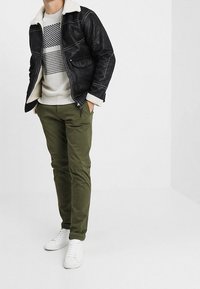 Schwarze Lederjacke mit cremefarbener Lammfellkragen, grauer gemusterter Pullover, olivgrüne Hose und weiße Sneakers, getragen von einer stehenden Person.
