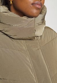 Chaqueta tipo plumón de suave tejido beige, con un cuello alto y detalles acolchados. Textura suave con un ligero brillo y hardware mínimo.