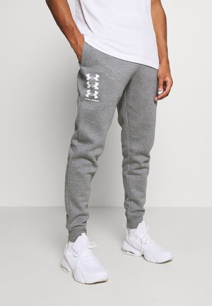 Man draagt grijze Under Armour joggingbroek met wit logo, witte sneakers en een wit T-shirt, staand tegen een effen achtergrond.