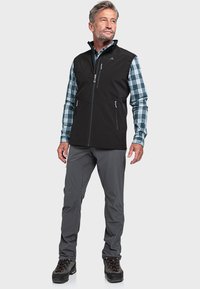 Schwarze Softshell-Weste mit hohem Kragen, Taschen mit Reißverschluss vorne, getragen über einem blau-grün karierten Hemd, kombiniert mit grauen Hosen und schwarzem Schuhwerk.