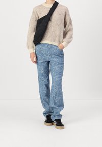 Hellbeiger Strickpullover mit strukturiertem Muster, blaue Hose mit Blumenmuster und schwarze Umhängetasche. Schwarze Sneaker mit weißen Akzenten.